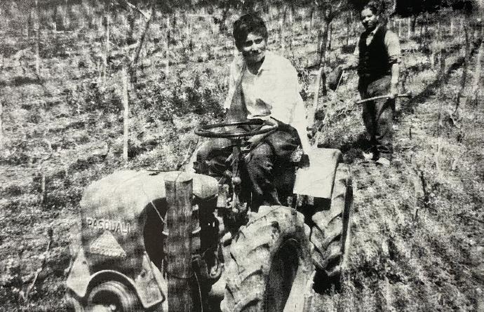 Fotografia storica in bianco e nero: un uomo alla guida di un trattore tra i campi e un altro in piedi accanto